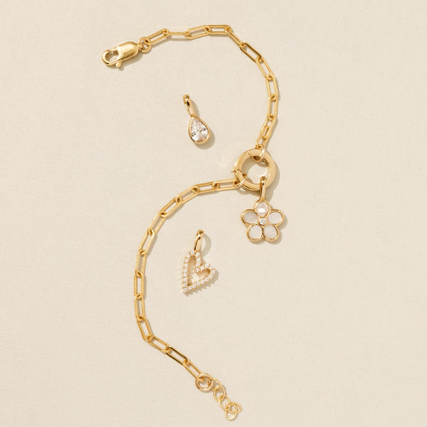 A gold paperclip chain charm bracelet with multiple gold charms, laying on a solid white surface.
