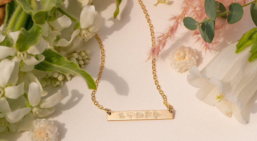 A gold horizontal bar necklace with engraved flowers, shown on a white surface surrounded by foliage and flowers.