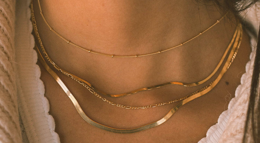 A close-up of a person's neck wearing four layered gold necklaces in different styles, two flat chains in varying widths, a gold link chain, and a gold chain with round gold stationary beads along it.