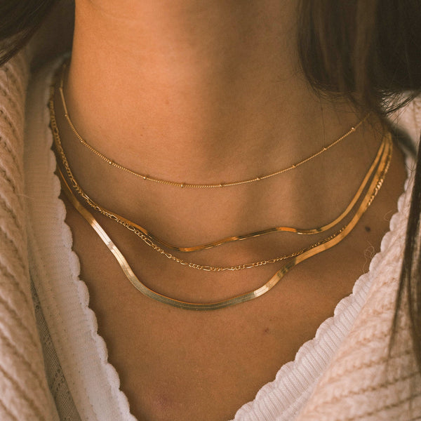 A close-up of a person's neck wearing four layered gold necklaces in different styles, two flat chains in varying widths, a gold link chain, and a gold chain with round gold stationary beads along it.