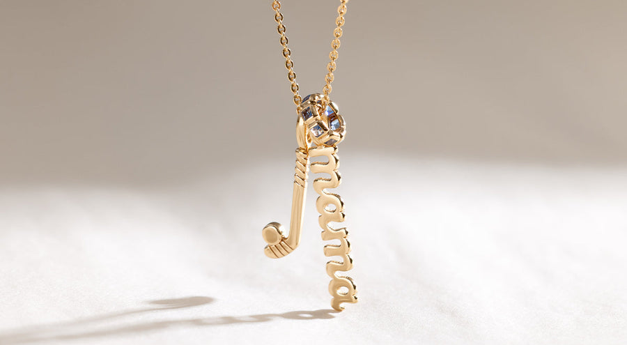 A delicate gold pendant chain necklace with a hockey stick charm, a script charm with the word 'mama', and a round ring charm with blue square gemstones.