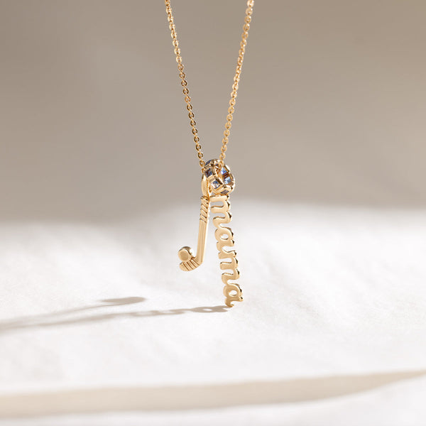 A delicate gold pendant chain necklace with a hockey stick charm, a script charm with the word 'mama', and a round ring charm with blue square gemstones.