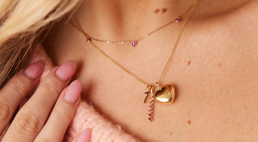 A person wearing a delicate gold necklace with stationary heart charms and a longer pendant necklace with multiple charms.