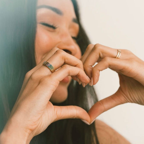 A woman with her hands in a heart shape, wearing multiple stacked rings with rainbow colored gemstones.