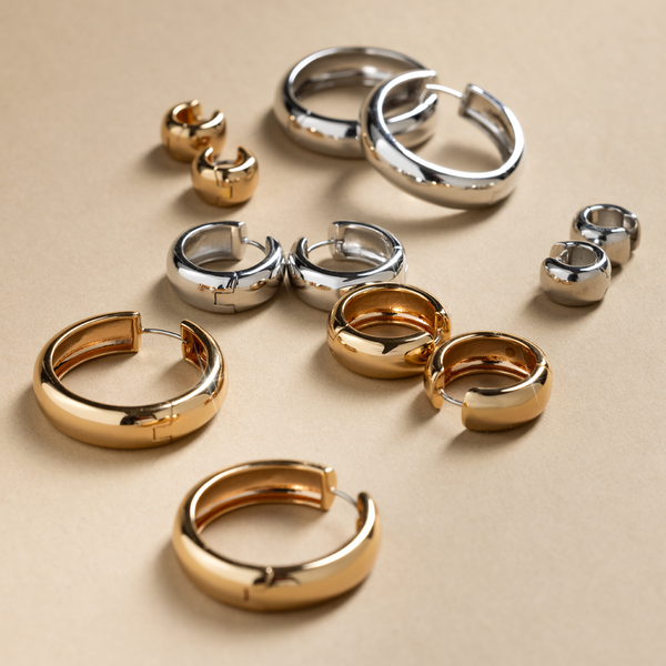 Multiple sizes of gold and silver chunky hoop earrings, laying scattered on a white surface with shadows.