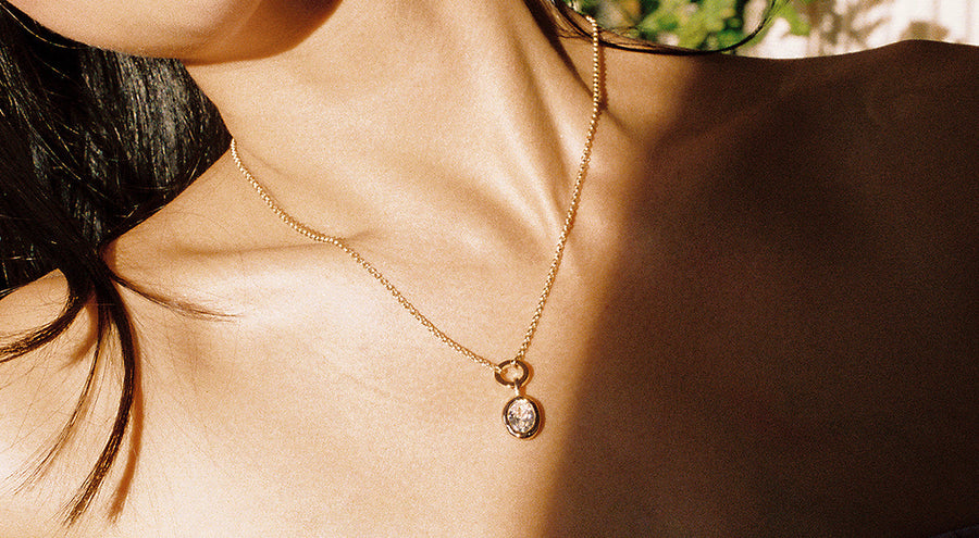 A woman in the sunlight wearing a gold charm necklace with a round ring in the center, holding a large clear oval gemstone charm.