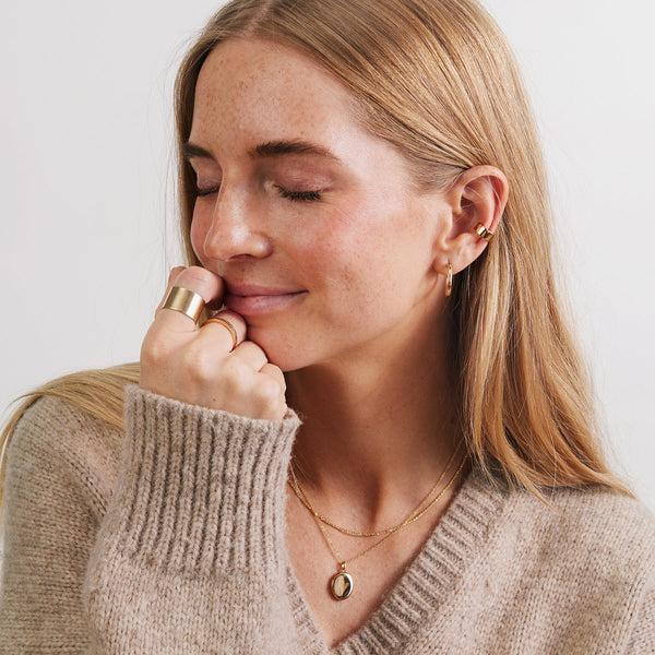 A woman in a light beige v-neck knit sweater wearing multiple gold rings, earrings, and a gold pendant necklace.