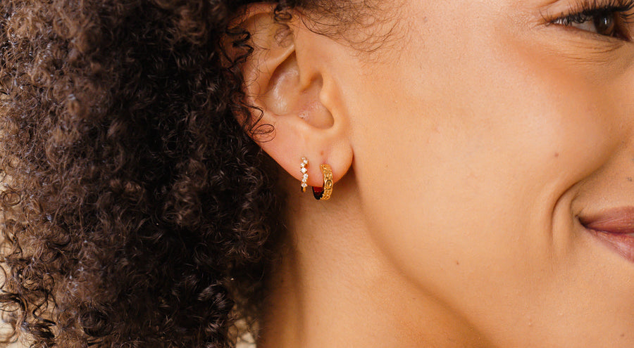 A close-up of a woman's ear wearing two pairs of gold huggie hoops, one covered with small clear accent stones.