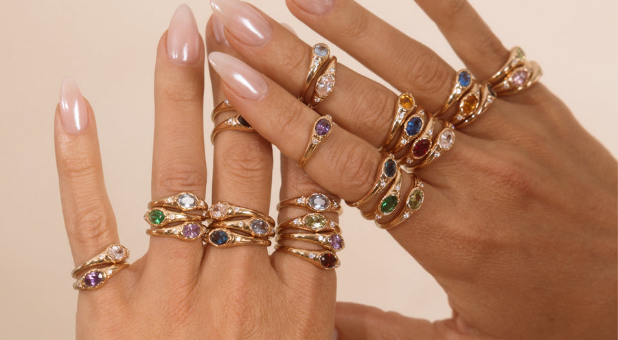 Hands wearing multiple gold signet rings with oval gemstones in varying colors.