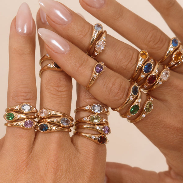 Hands wearing multiple gold signet rings with oval gemstones in varying colors.