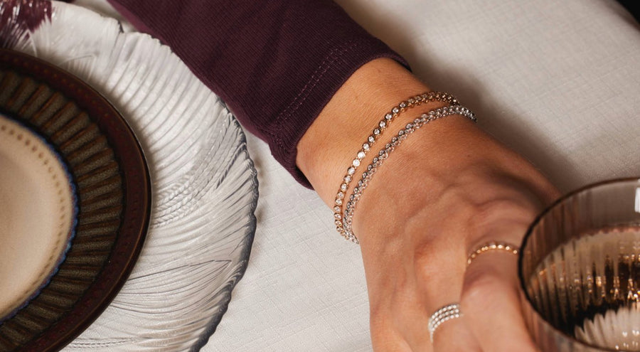 A woman's arm resting on a table holding a glass, wearing two tennis bracelets and matching rings