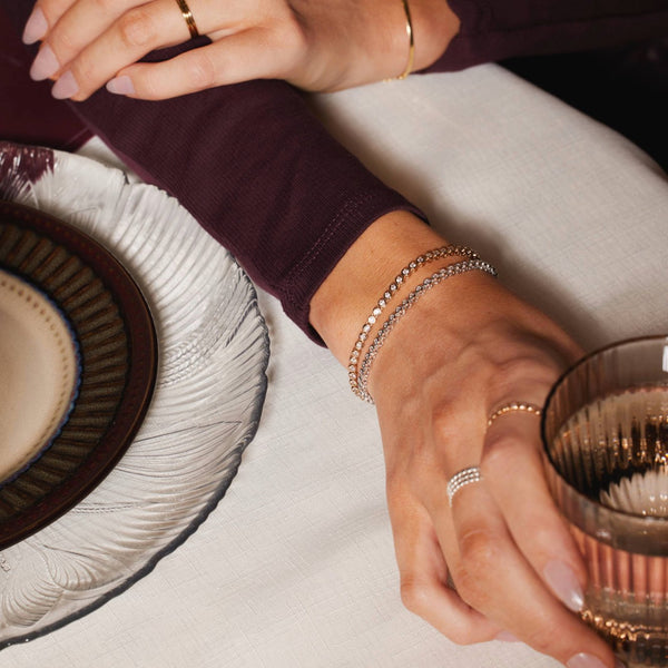 A woman's arm resting on a table holding a glass, wearing two tennis bracelets and matching rings