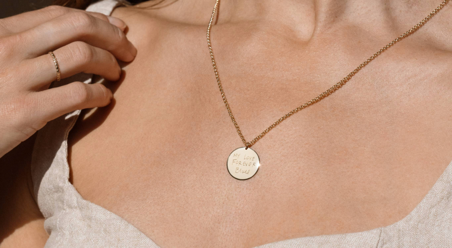 A lifestyle image of a person wearing a gold pendant necklace featuring a large round disc charm, engraved with text, paired with a gold ring and beige linen top.