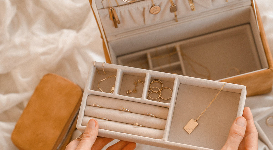 A person holding the insert to a rectangular brown leather jewelry box, sitting open with multiple pieces of gold jewelry inside.