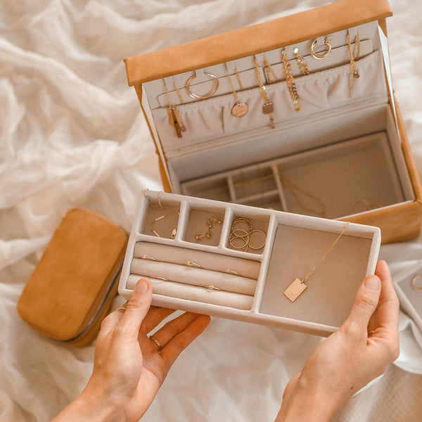 A person holding the insert to a rectangular brown leather jewelry box, sitting open with multiple pieces of gold jewelry inside.