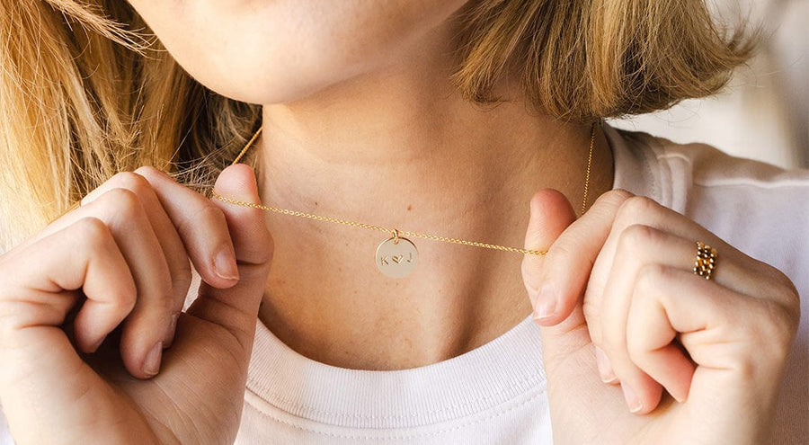 A woman in a white t-shirt holding out a gold pendant chain necklace with a round engraved disc charm.