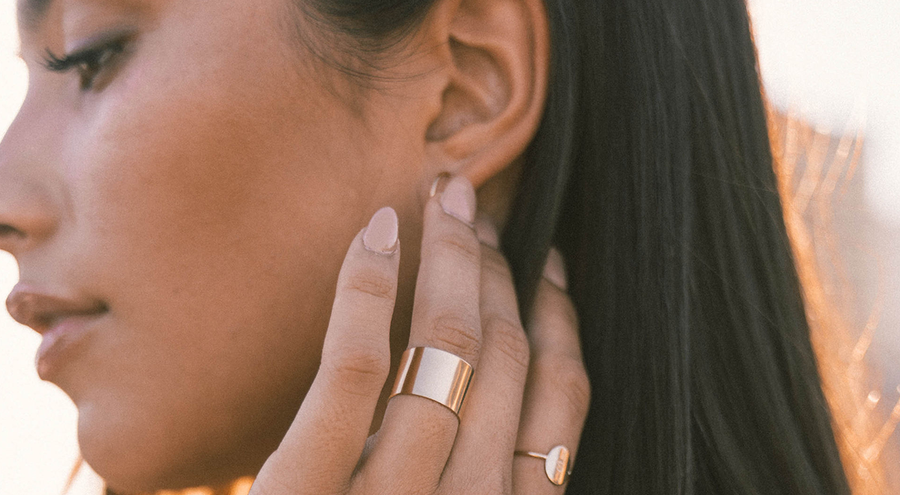 A person's hand against their cheek wearing a thick gold cigar band ring and a dainty engraved oval ring.