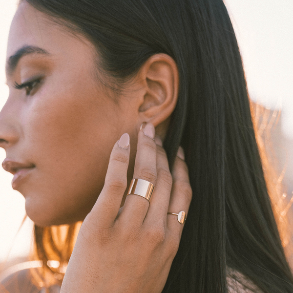 A person's hand against their cheek wearing a thick gold cigar band ring and a dainty engraved oval ring.