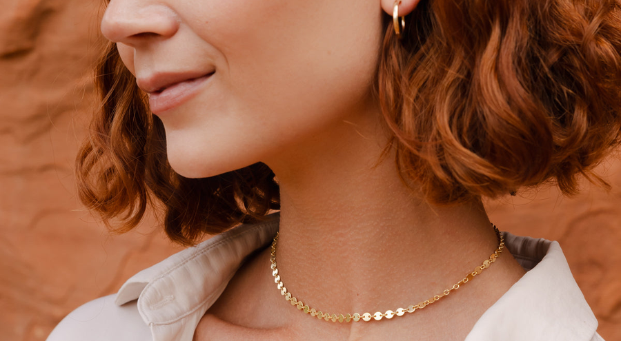 A red-haired woman in a white button up top standing in front of red rocks, wearing a gold choker necklace with flat round discs linked together.
