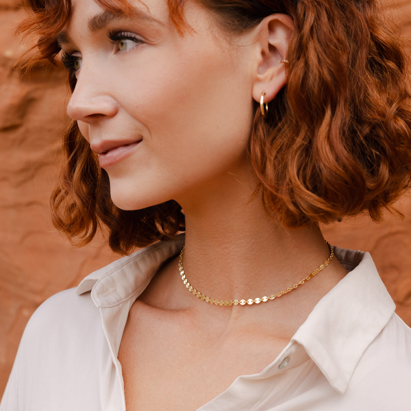 A red-haired woman in a white button up top standing in front of red rocks, wearing a gold choker necklace with flat round discs linked together.