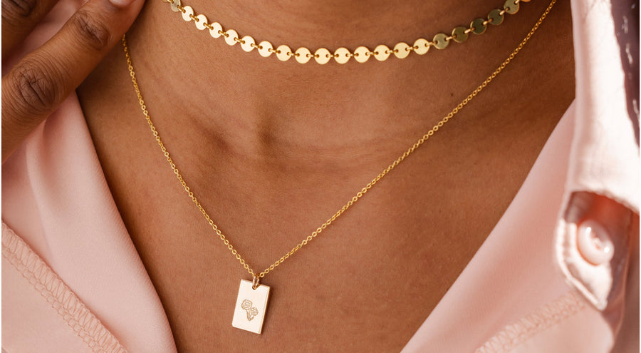 A woman wearing a gold choker necklace made up of small flat round discs, and a longer pendant chain with a flat polished engraved rectangle charm.