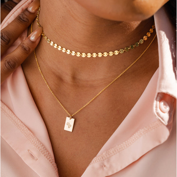 A woman wearing a gold choker necklace made up of small flat round discs, and a longer pendant chain with a flat polished engraved rectangle charm.