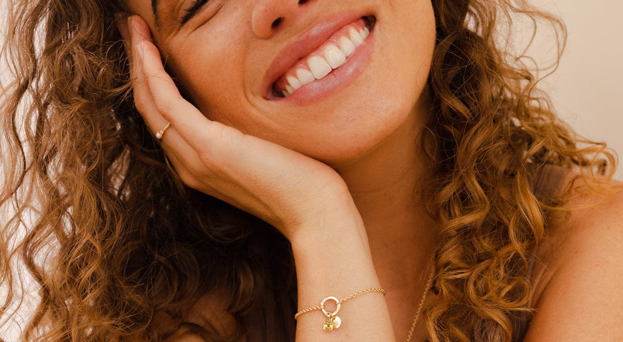 A smiling woman wearing a gold chain charm bracelet with multiple charms.