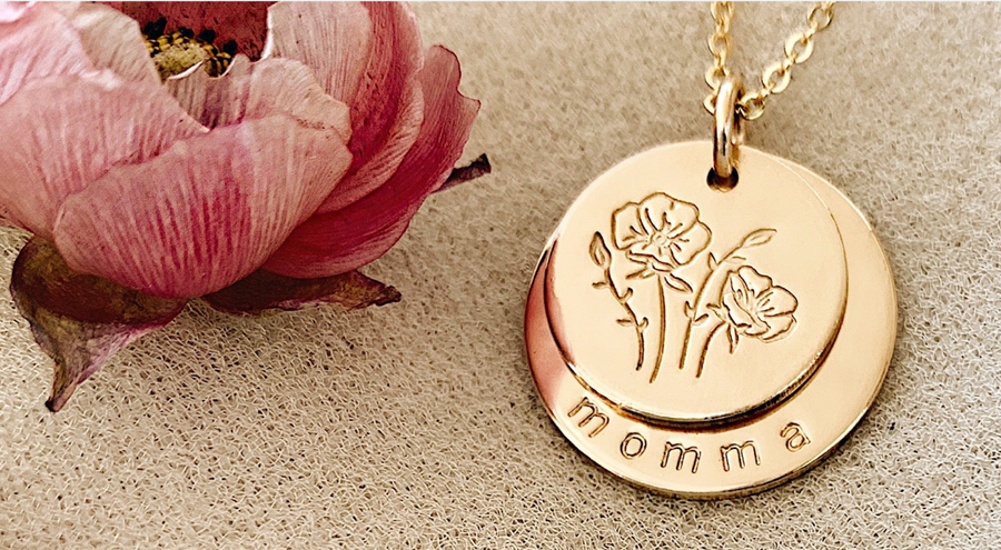 A pendant chain with two layered gold discs, the top one with a floral design and the bottom with the word 'momma' along the bottom edge.