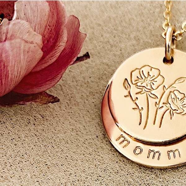 A pendant chain with two layered gold discs, the top one with a floral design and the bottom with the word 'momma' along the bottom edge.