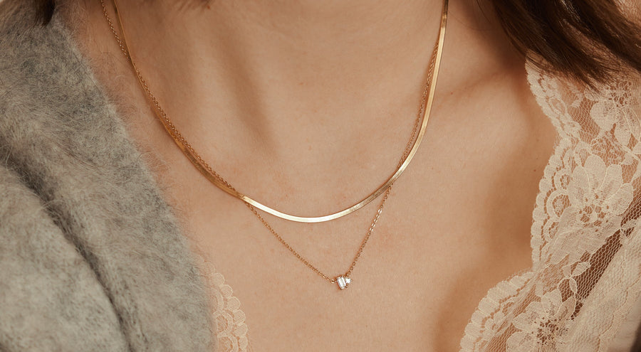 A woman wearing a cream lace top and grey knit cardigan, showcasing a thick gold chain necklace layered with a delicate gold necklace with a stationary clear baguette and teardrop gemstone charm.