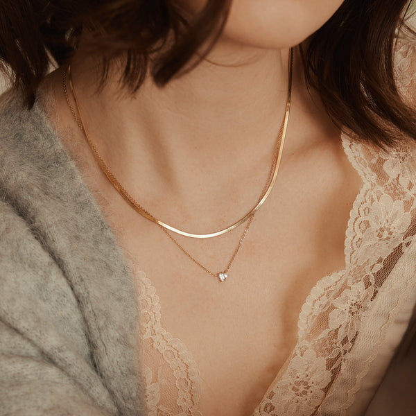 A woman wearing a cream lace top and grey knit cardigan, showcasing a thick gold chain necklace layered with a delicate gold necklace with a stationary clear baguette and teardrop gemstone charm.
