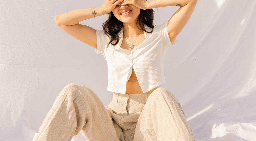 A woman in a white shirt and khaki pants covering her eyes, sitting with her knees up on a white linen background.
