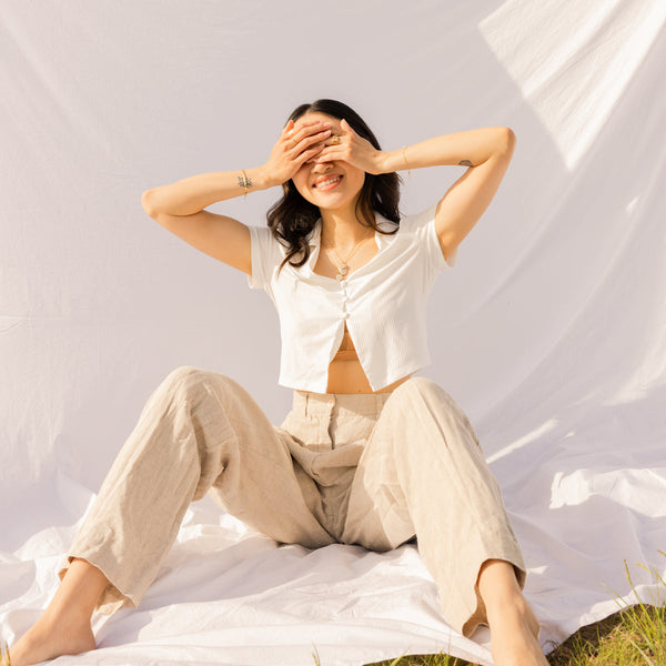 A woman in a white shirt and khaki pants covering her eyes, sitting with her knees up on a white linen background.