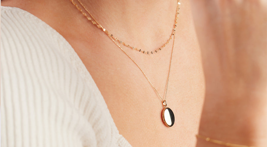 A person wearing a delicate textured gold chain necklace layered with a gold oval locket pendant necklace and a white ribbed knit shirt.