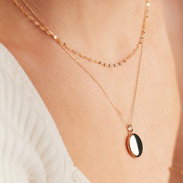 A person wearing a delicate textured gold chain necklace layered with a gold oval locket pendant necklace and a white ribbed knit shirt.