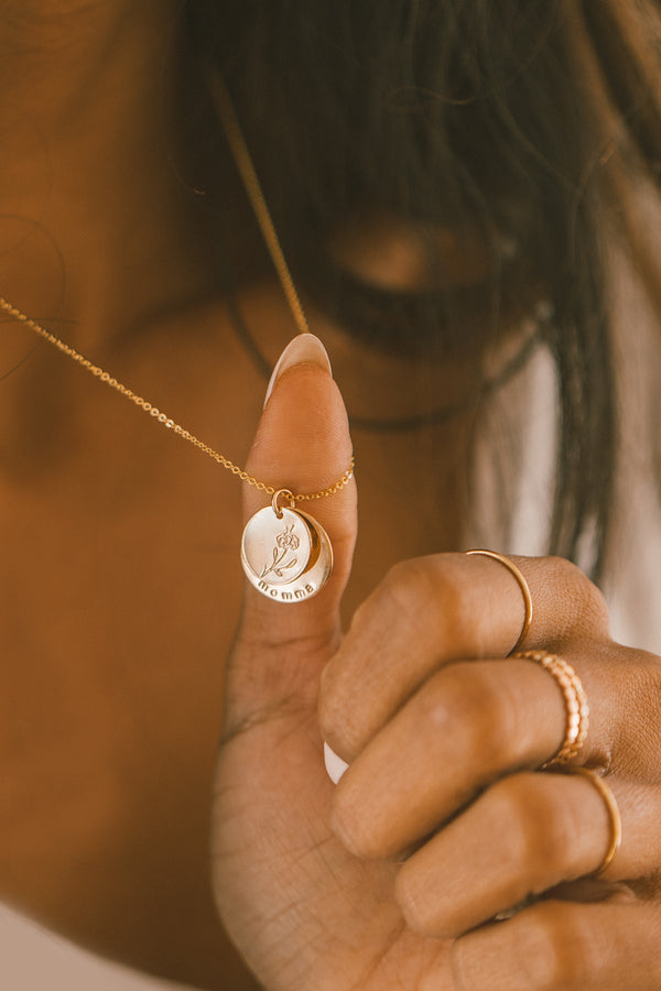 A close up lifestyle photo of a person pulling a dainty gold pendant chain on their neck, showing two, round layered discs. The top disc shows a floral motif, and the larger bottom disc has the word 'momma' across the bottom. The hand also has multiple gold rings.