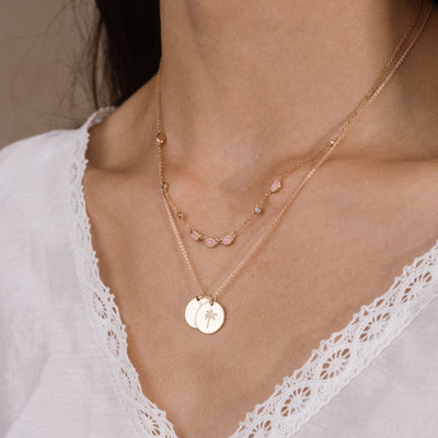 A close-up of a person's neck and chest wearing a layered necklace set. The jewelry features two delicate gold chains: one with small oval-shaped pink gemstones, and another with two small circular gold pendants engraved with a palm tree and a heart.