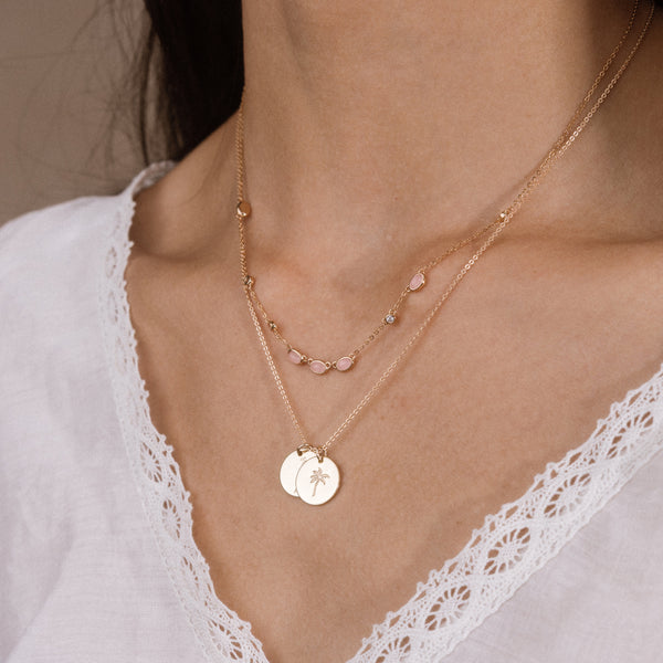 A close-up of a person's neck and chest wearing a layered necklace set. The jewelry features two delicate gold chains: one with small oval-shaped pink gemstones, and another with two small circular gold pendants engraved with a palm tree and a heart.