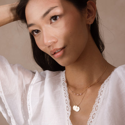 A close-up lifestyle shot of a woman wearing layered gold necklaces, including a round disc pendant and a delicate chain with a small pink oval stones, paired with a white lace-trimmed blouse against a soft beige background.