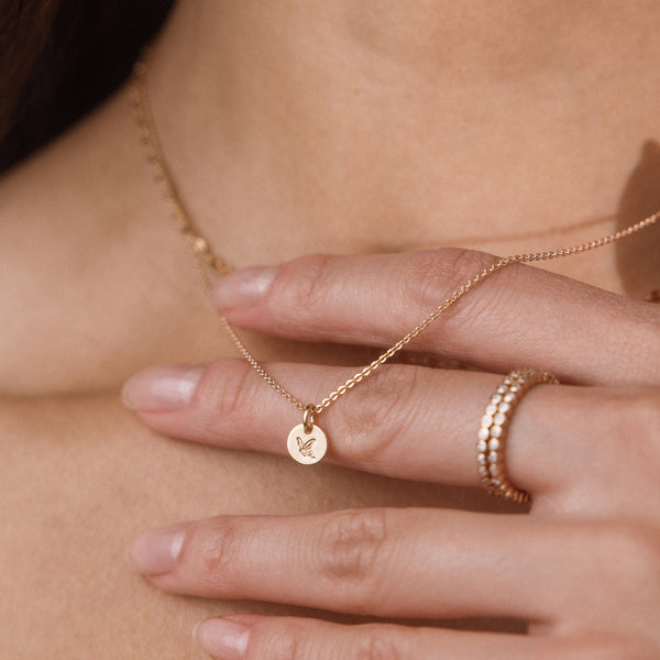 A close-up lifestyle shot of a model wearing a delicate gold chain necklace with a small circular pendant engraved with a butterfly, paired with two stacked gold rings.