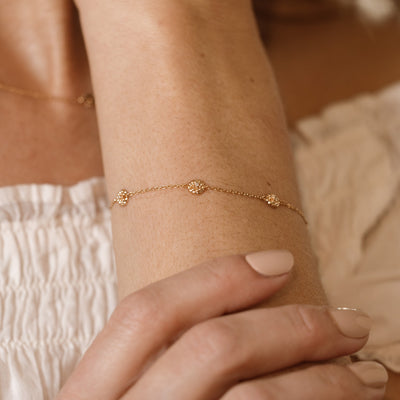 A lifestyle image of a dainty gold bracelet on a person's wrist with stationary textured marigold flowers with small accent stones in the center along the chain.