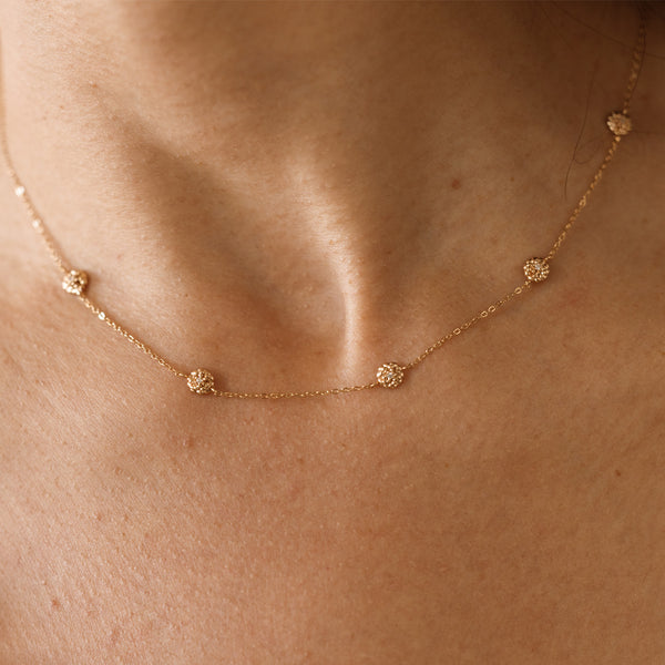 A lifestyle image of a dainty gold necklace on a person's neck with stationary textured marigold flowers with small accent stones in the center along the chain.