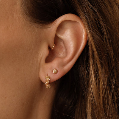 A close up lifestyle shot of a person's ear wearing a gold hoop earring with marigold flowers and clear accent stones, and a gold stud earring with one singular marigold flower and a single accent stone.