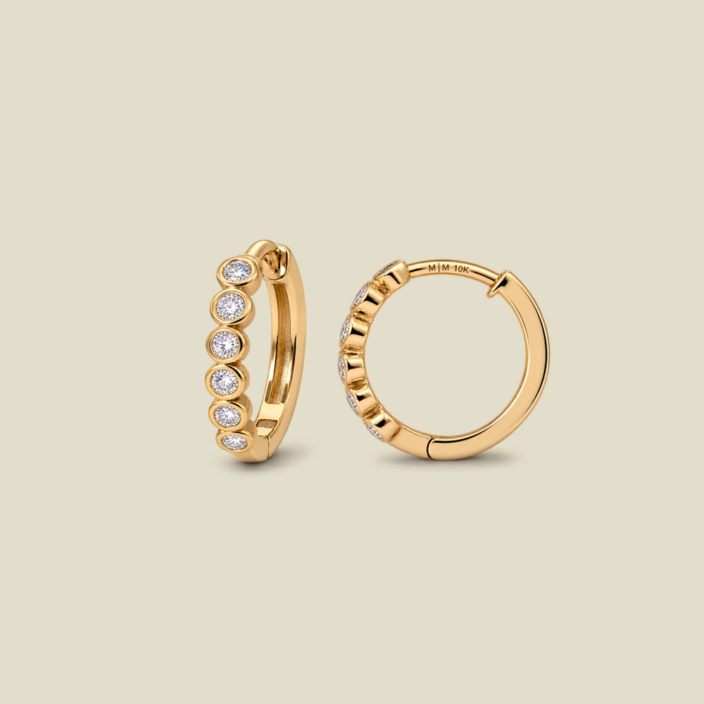 A pair of gold huggie hoop earrings, one shown from the front with six bezel-set round clear diamonds and the other from the side showing the hinge closure. The earrings are displayed on a solid light beige background.