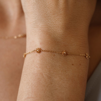 A lifestyle image of a dainty gold bracelet on a person's wrist with stationary textured chrysanthemum flowers with small accent stones in the center along the chain.
