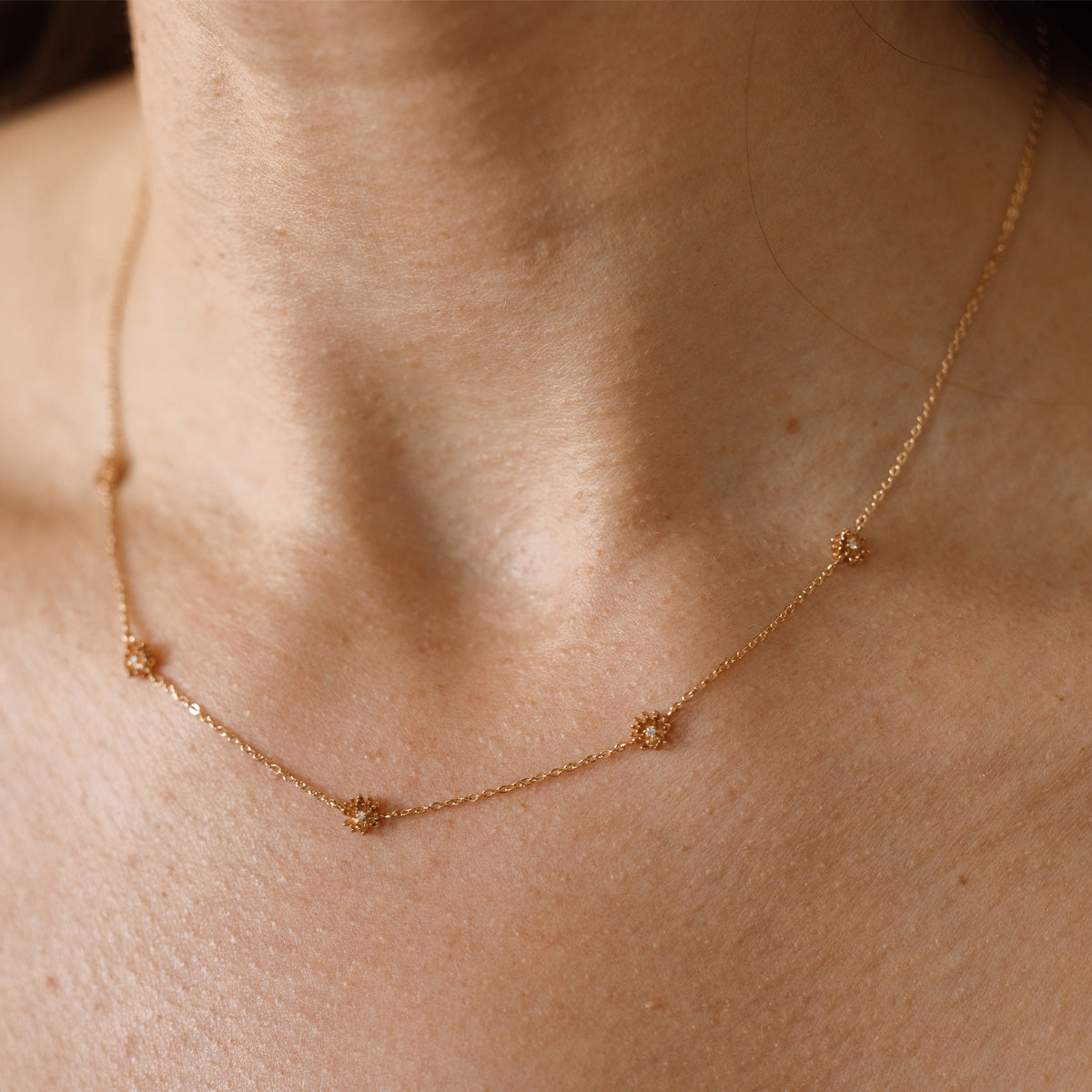 A lifestyle image of a dainty gold necklace on a person's neck with stationary textured chrysanthemum flowers with small accent stones in the center along the chain.