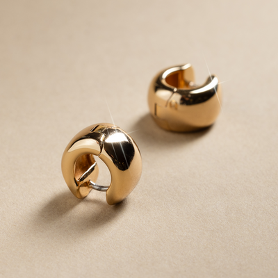 A wide yellow-gold huggie hoop earring, displayed on a textured beige background.