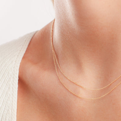 A close up shot of a person's neck wearing two gold delicate chains layered, with a white knit top.