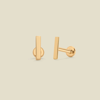 A pair of yellow-gold bar stud earrings with flat backings, set against a solid beige background.