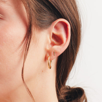 A close up photo of a person's ear wearing a polished gold hoop and a single gold bar stud earring.